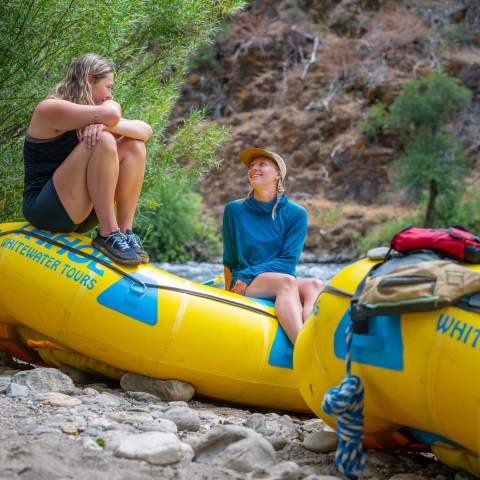 Two people sitting on yellow rafts by a riverbank, surrounded by greenery.