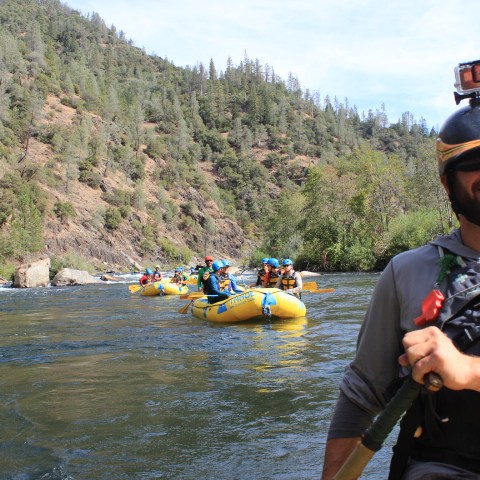 Smiling man in helmet leads group rafting on river with forested hills.