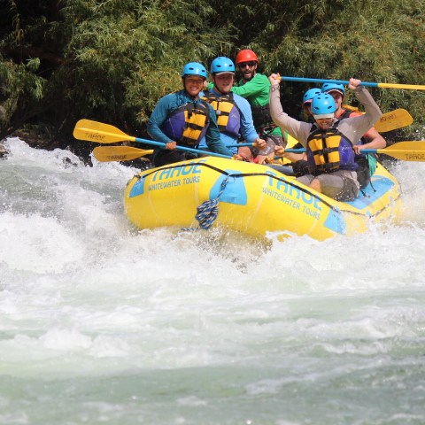 Group of people wearing helmets white-water rafting in a yellow raft with paddles.