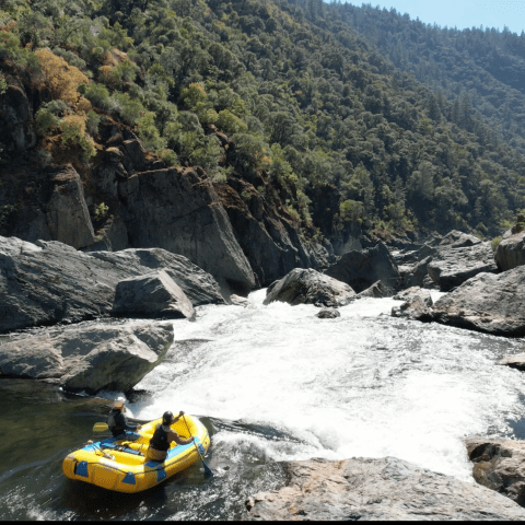Two people rafting on a river surrounded by rocky terrain and lush trees.