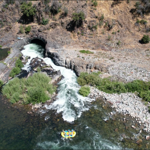 River with a raft going through rapids beside rocky terrain and tunnel.