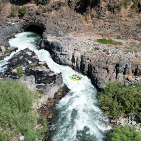 Rafting group navigating rapids near rocky terrain and a tunnel entrance.