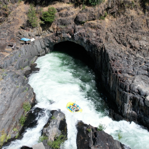 Rafting group navigating whitewater rapids near a rocky cave entrance.