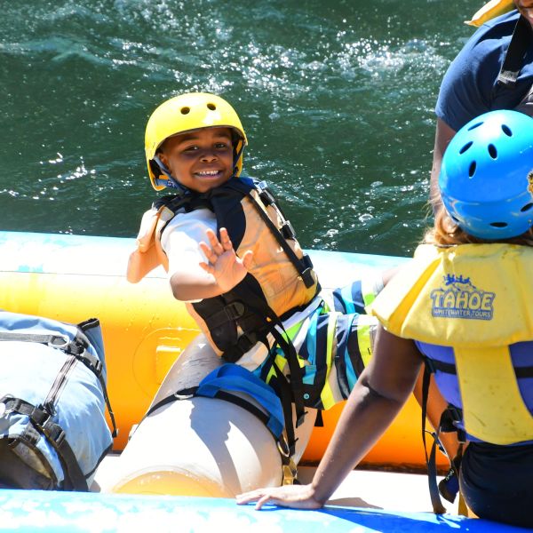 Children in helmets and vests rafting on a river, smiling and waving.