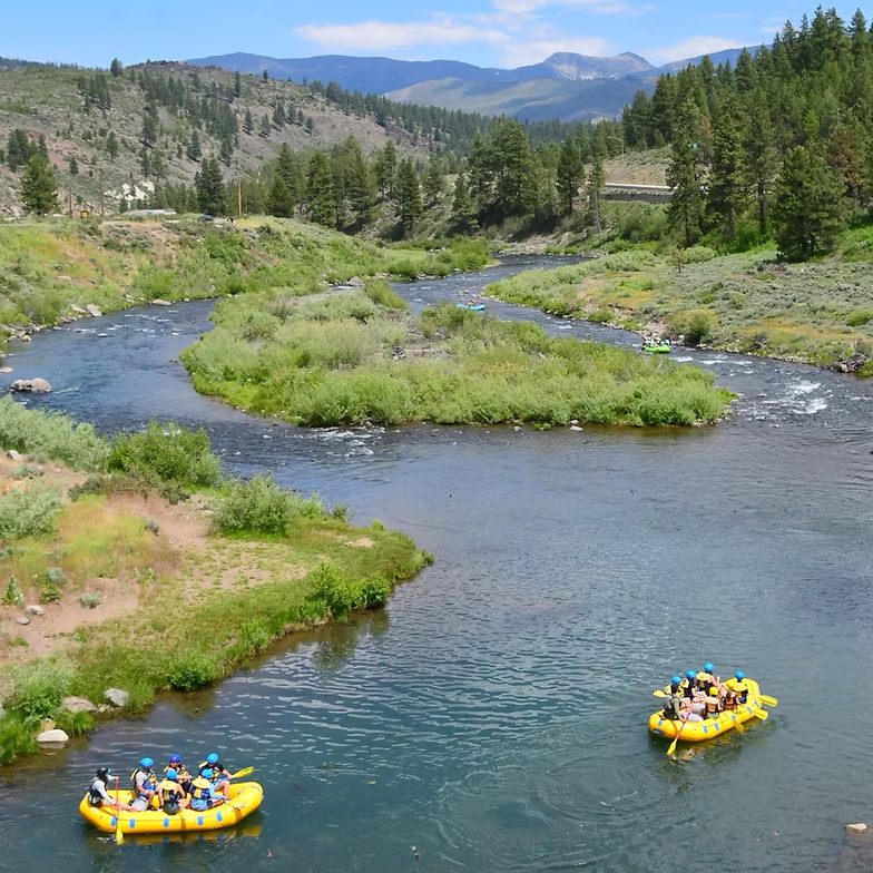 Two rafts with people on a river, surrounded by forested hills and a clear sky.