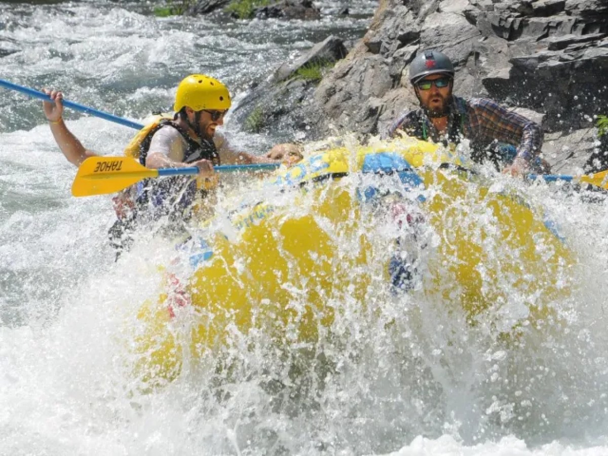 Three people white water rafting in rough river, wearing helmets and life jackets.