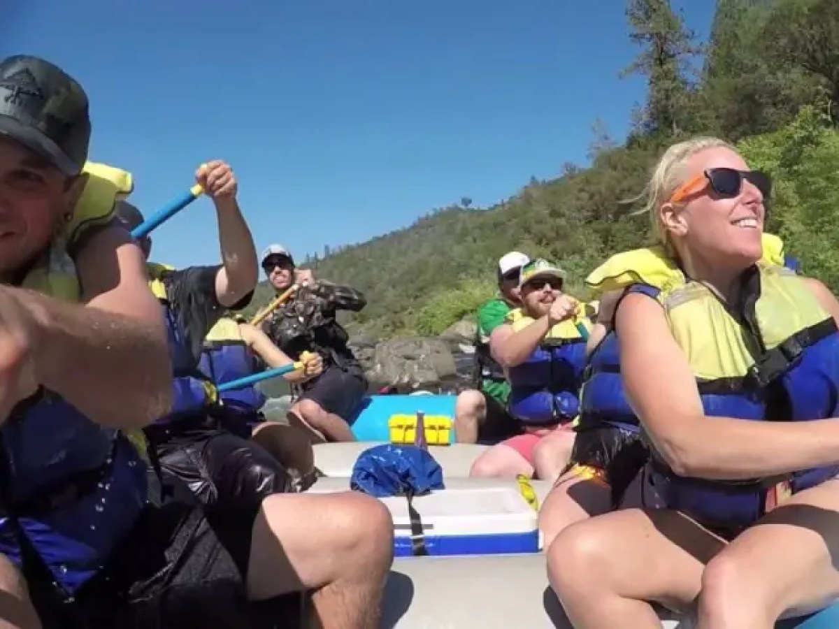 Group of people rafting on a river, wearing life vests and paddling, with trees in the background.